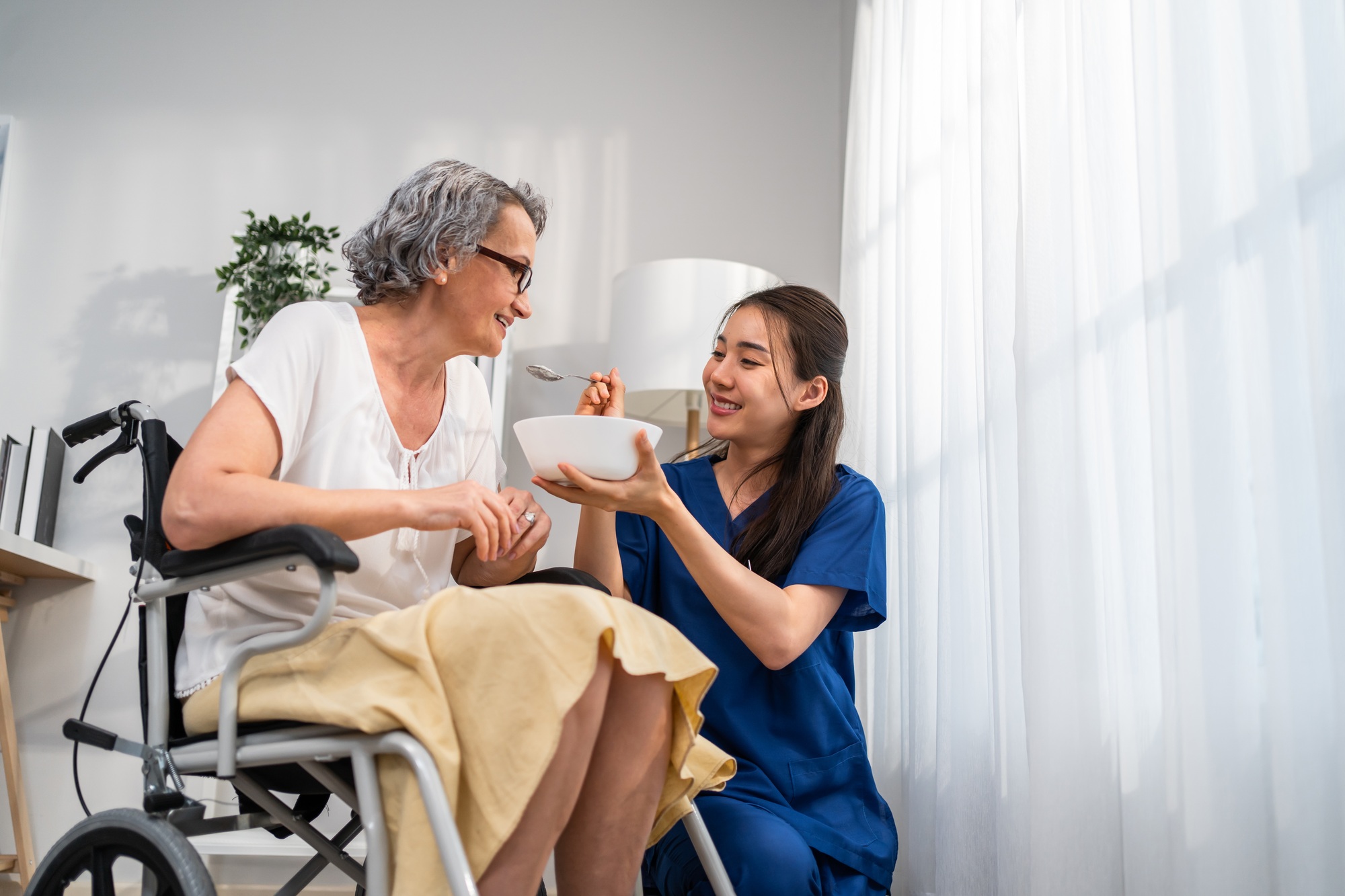 Asian caregiver nurse serve food to Caucasian senior older woman at home.