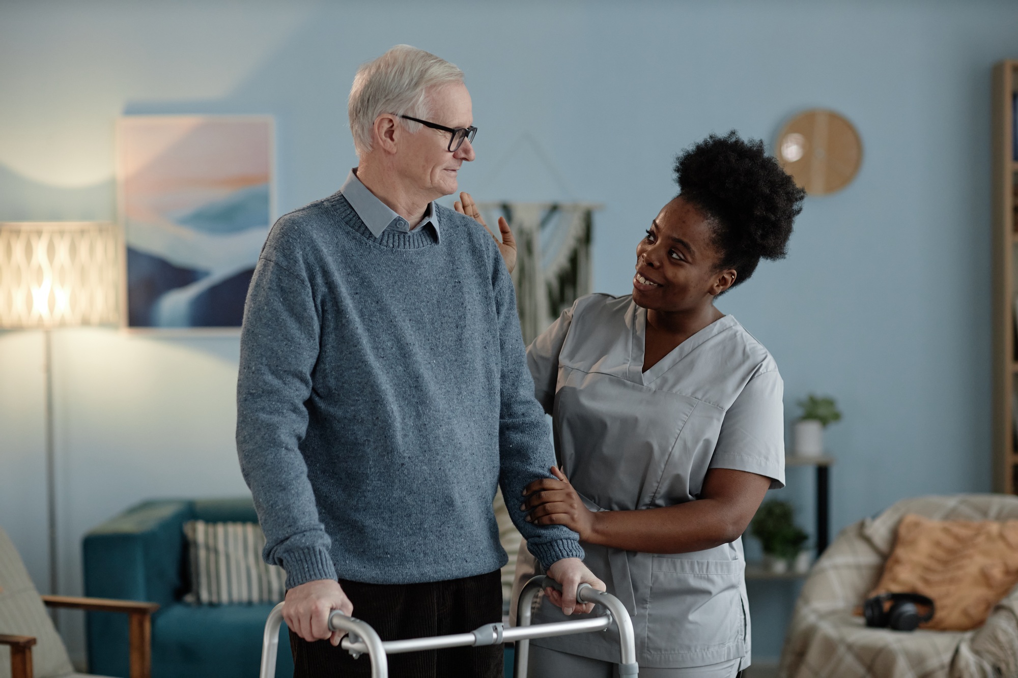 Positive Black Female Caregiver Helping Senior Man Walking with Frame Indoors
