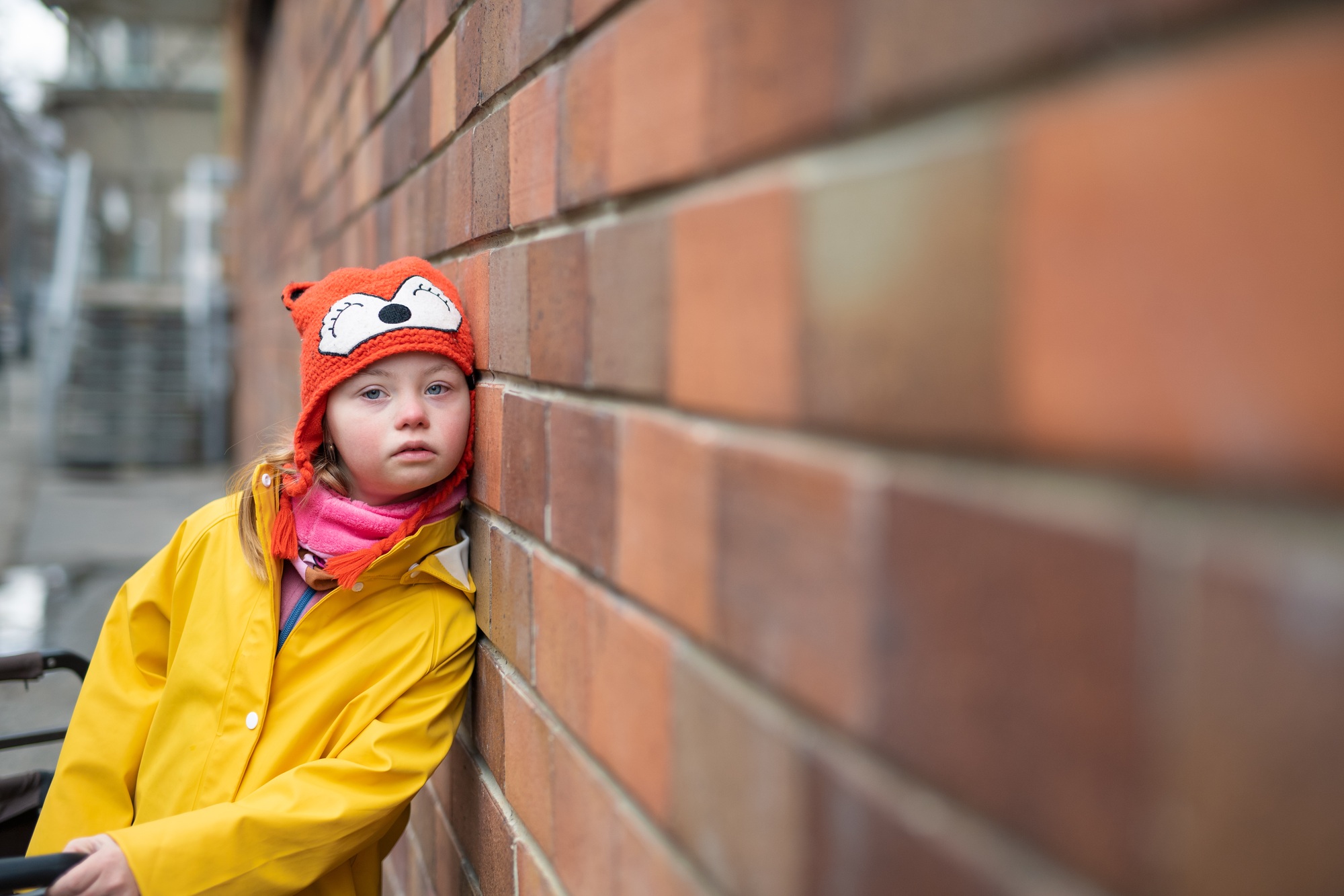 Little girl with Down syndrome leaning against brick wall in winter.